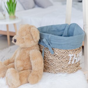 May include: A light brown teddy bear sits on a white fluffy rug next to a woven basket with a blue linen liner. The basket has the word "Thomas" embroidered on the side.