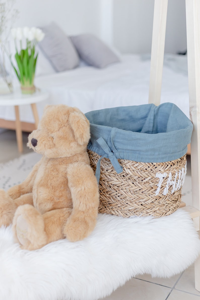 May include: A light brown teddy bear sits on a white fluffy rug next to a woven basket with a blue linen liner. The basket has the word "Thomas" embroidered on the side.