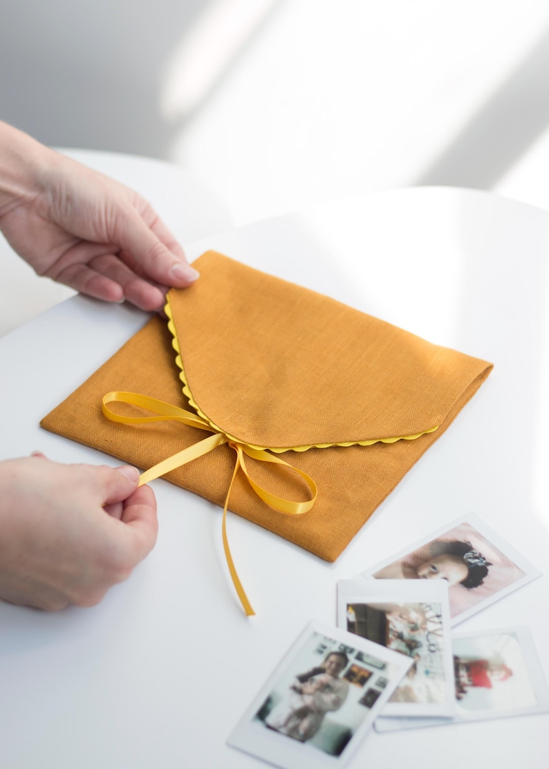 May include: A mustard yellow fabric envelope with a scalloped edge and a yellow ribbon tied in a bow. The envelope is open and there are several instant photos inside.