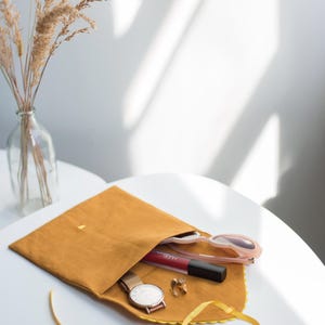 May include: A mustard yellow fabric pouch with a yellow ribbon. The pouch is open and contains a watch, a lipstick, sunglasses, and earrings.