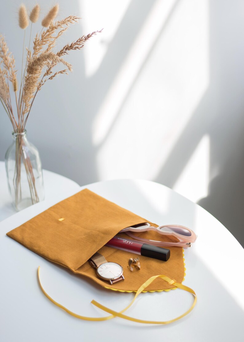 May include: A mustard yellow fabric pouch with a yellow ribbon. The pouch is open and contains a watch, a lipstick, sunglasses, and earrings.