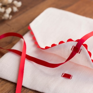 May include: A white linen envelope with a red ribbon tied around it. The envelope has a scalloped edge and is decorated with red stitching.