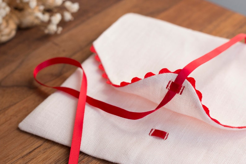 May include: A white linen envelope with a red ribbon tied around it. The envelope has a scalloped edge and is decorated with red stitching.