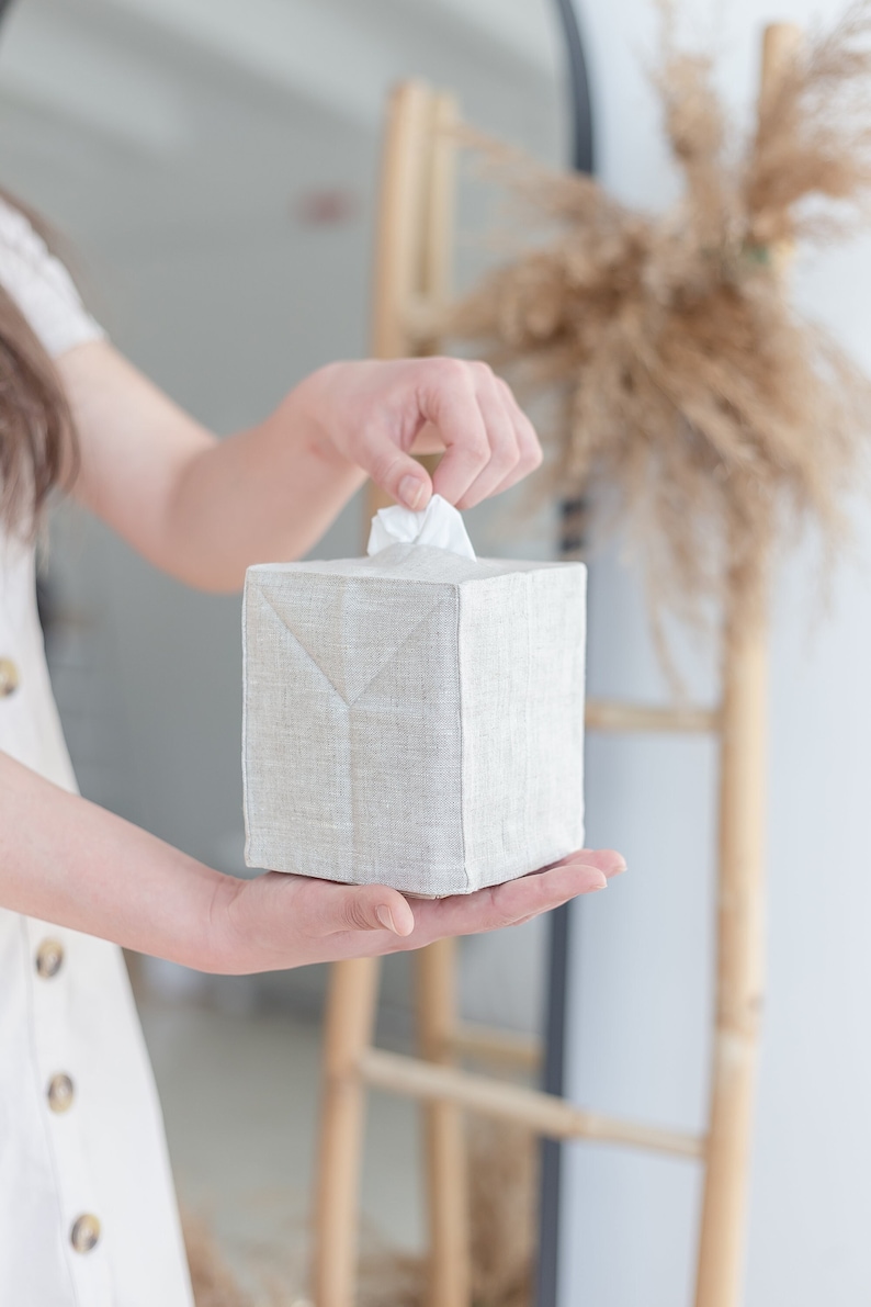 May include: A square tissue box cover made of light beige, textured fabric. The cover features a clean, structured design with visible seams along the edges and a central opening at the top from which a single white tissue is being pulled.