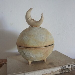 Könnte beinhalten: Keramik-Schmuckkästchen mit kugelförmigem Körper und drei kleinen Füßen. Der Deckel ist mit einem Halbmond mit lächelndem Gesicht gekrönt. Die Box hat eine strukturierte, cremefarbene Oberfläche und steht auf einem Stapel Bücher.