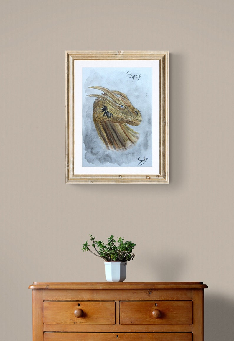May include: A framed watercolor painting of a dragon's head, featuring shades of gold and brown. The dragon's eye is blue, and the artwork is signed. The word "Syrax" is written above the dragon's head. The frame is gold.