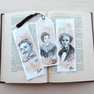 May include: Three watercolor bookmarks featuring portraits of historical figures, resting on an open book. Each bookmark has a brown tassel. The portraits are in black and white with a sepia-toned background. The names of the figures are written below the portraits.