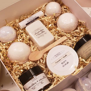 May include: A gift box filled with lavender and vanilla scented bath products. The box contains bath bombs, soap, body scrub, body cream, lip balm, and a wooden spoon. The products are arranged on gold-colored paper filler.