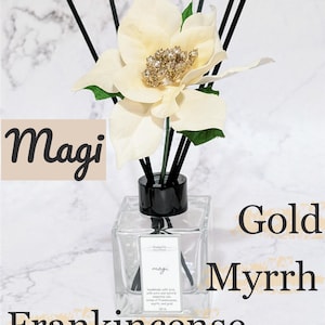 May include: A clear glass diffuser bottle with black reeds and a cream-colored flower with a gold center. The bottle is labeled "Magi" and the words "Gold Myrrh Frankincense" are also visible.
