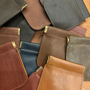 May include: A collection of leather coin purses in various colors, including brown, tan, and navy. Each purse has a rounded shape, stitched edges, and a gold-colored metal clasp. The purses are arranged on a wooden surface.