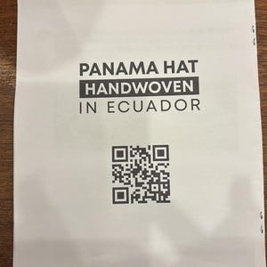 Puede incluir: Una etiqueta de papel blanca con texto negro que dice "Panama Hat Handwoven in Ecuador". Un c&oacute;digo QR en blanco y negro est&aacute; debajo del texto.