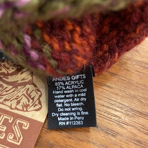 Op de afbeelding: Een zwart label met witte tekst dat "ANDES GIFTS 83% ACRYLIC 17% ALPACA Handwas in koud water met een mild wasmiddel. Drogen aan de lucht plat. Geen bleekmiddel. Niet wringen. Stomen is mogelijk. Gemaakt in Peru RN #112363" aangeeft