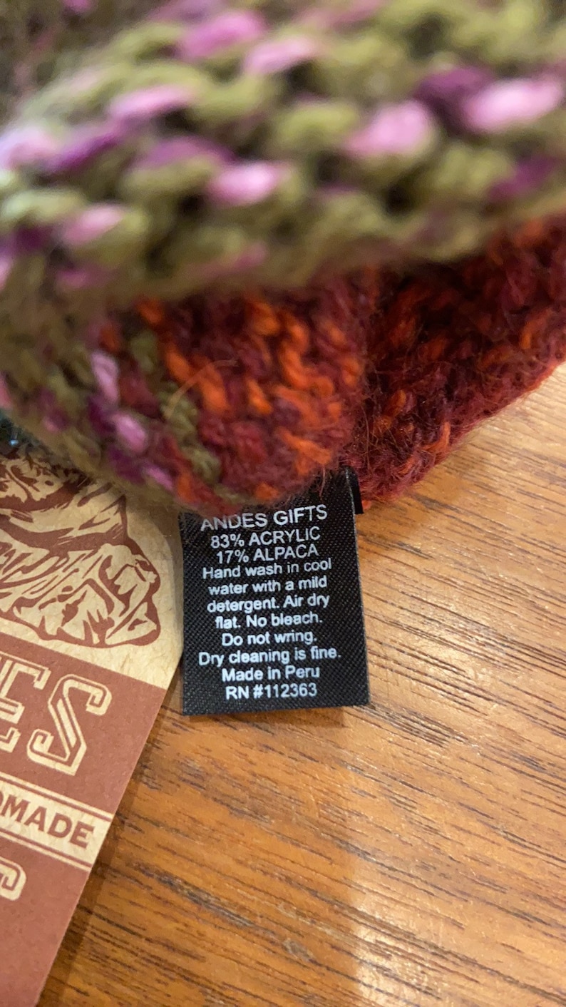 Op de afbeelding: Een zwart label met witte tekst dat "ANDES GIFTS 83% ACRYLIC 17% ALPACA Handwas in koud water met een mild wasmiddel. Drogen aan de lucht plat. Geen bleekmiddel. Niet wringen. Stomen is mogelijk. Gemaakt in Peru RN #112363" aangeeft