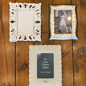 May include: Three decorative picture frames on a wooden surface. One frame is white with ornate carvings, another is silver with a photo of a bride, and the third is white with the text "The Lenox Portrait Gallery".
