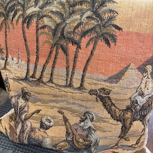 May include: A square throw pillow featuring a woven tapestry design of a desert landscape. The scene depicts palm trees, pyramids, and figures in traditional attire, including a person riding a camel and another playing a stringed instrument.
