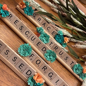 May include: Five wooden tiles with the words "Blessed", "Rescued", "Forgiven", "Trust", and "Grace" spelled out. Each tile is decorated with small, colorful paper flowers.