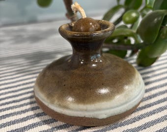 May include: A small, round, ceramic oil lamp with a glossy brown glaze on the top and a matte, unglazed terracotta base. The lamp features a flared neck and a central opening containing a single, light-colored cotton wick for illumination.