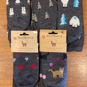 May include: Four pairs of alpaca bamboo casual socks with different designs. The socks are gray with colorful patterns. The designs include snowmen, trees, hearts, cats, and a llama. The socks are packaged in brown cardboard with the brand name "RedMaple" printed on them.