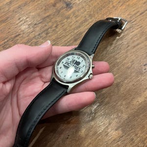 May include: A wristwatch with a white face, silver casing, and a black leather band. The watch face displays the word "PICRES" and the numbers 1 through 12. The watch is held against a wooden surface.