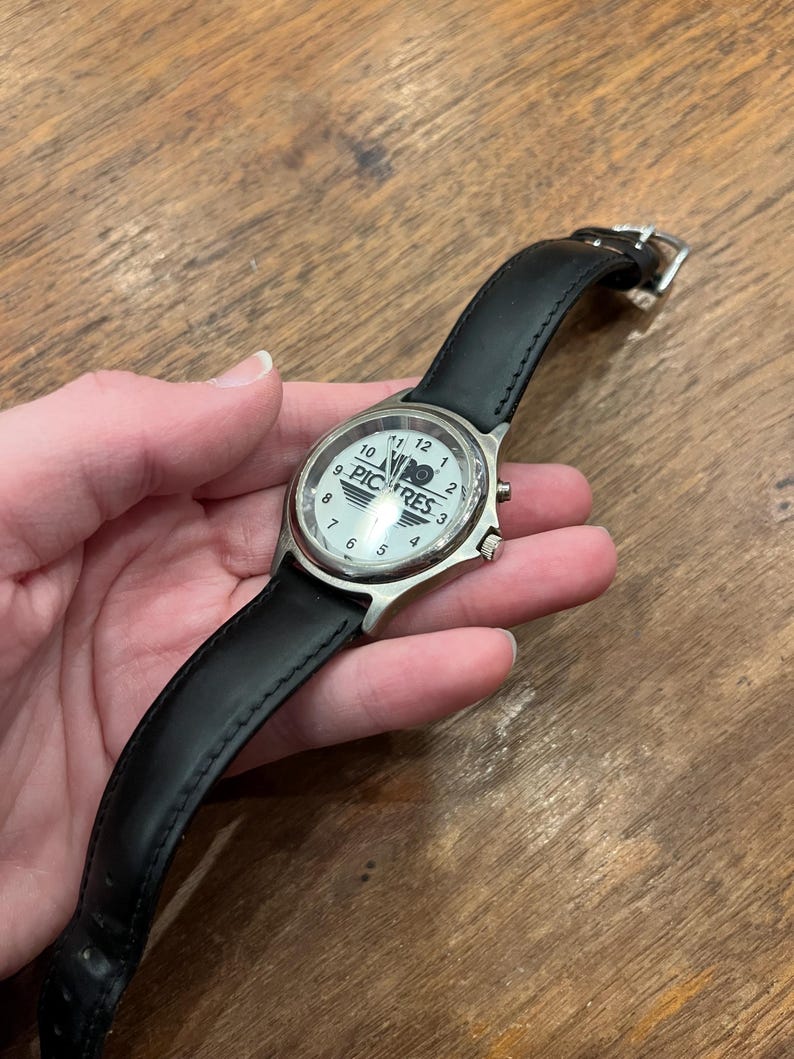 May include: A wristwatch with a white face, silver casing, and a black leather band. The watch face displays the word "PICRES" and the numbers 1 through 12. The watch is held against a wooden surface.