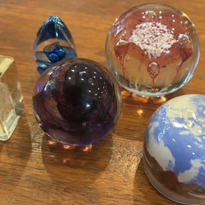 May include: A collection of five glass paperweights including a clear rectangular block with an internal etching, a small blue teardrop shape, a dark purple sphere, a clear sphere with red and white inclusions, and a blue and white earth-like sphere.
