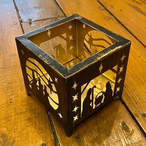 以下が含まれることがあります： 金色の仕上げが施された黒い金属製のキャンドルホルダー。ホルダーは立方体形で、星がついたキリスト降誕の場面が切り抜かれたデザインになっています。
