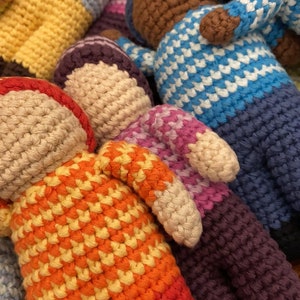 May include: A group of colorful crocheted dolls with different patterns and colors. The dolls are made with a variety of yarn colors, including red, orange, yellow, blue, and purple. The dolls are all in a pile, and some of them are partially obscured by others.