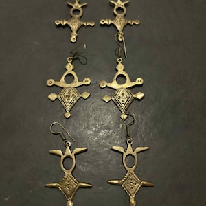 May include: Six pairs of brass-colored earrings with a cross-like design. Each earring features a diamond-shaped center with intricate patterns and a hook for the ear. The earrings are laid out on a dark surface.