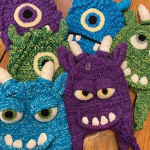 May include: Five colorful crocheted monster hats with different facial expressions. The hats are made with a variety of textures and colors, including purple, blue, green, and white.