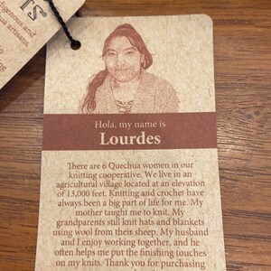 Op de afbeelding: Een bruin label met een foto van een vrouw en de tekst "Hola, my name is Lourdes." De tekst gaat verder met "There are 6 Quechua women in our knitting cooperative. We live in an agricultural village located at an elevation of 13,000 feet. Knitting and crochet have always been a big part of life for me. My mother taught me to knit. My grandparents still knit hats and blankets using wool from their sheep. My husband and I enjoy working together, and he often helps me put the finishing touches on my knits. Thank you for purchasing our products! Learn more about our cooperatives at WWW.ANDESGIFTS.COM"