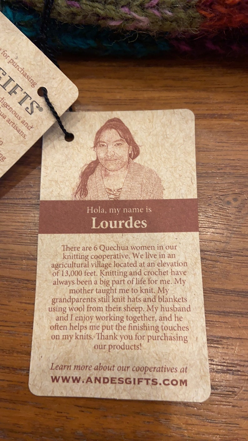 Op de afbeelding: Een bruin label met een foto van een vrouw en de tekst "Hola, my name is Lourdes." De tekst gaat verder met "There are 6 Quechua women in our knitting cooperative. We live in an agricultural village located at an elevation of 13,000 feet. Knitting and crochet have always been a big part of life for me. My mother taught me to knit. My grandparents still knit hats and blankets using wool from their sheep. My husband and I enjoy working together, and he often helps me put the finishing touches on my knits. Thank you for purchasing our products! Learn more about our cooperatives at WWW.ANDESGIFTS.COM"