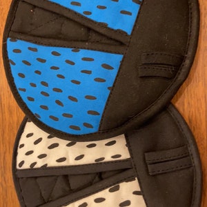 May include: Two round black pot holders with a blue and white patterned design. The pot holders have a black fabric border and a black fabric strap.