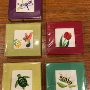 May include: Five small square frames with different colored backgrounds and nature-themed images inside. The frames are made of a light brown material and have a linen-like texture. The images include a dragonfly, a hummingbird, a tulip, a turtle, and a caterpillar.