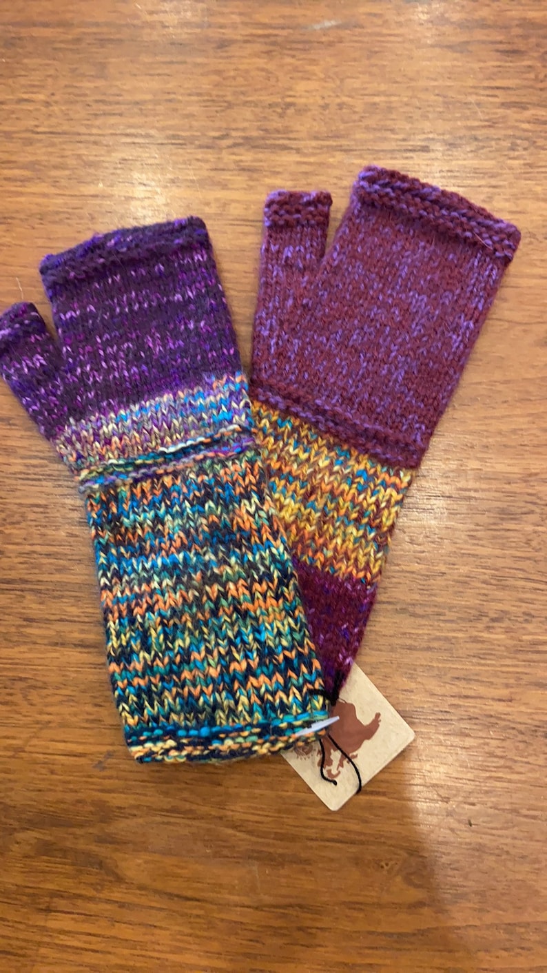Op de afbeelding: Een paar vingerloze handschoenen in een kleurenschema van paars, oranje en blauw. De handschoenen zijn gebreid en hebben een textuurpatroon. E&eacute;n handschoen is effen paars met een streep oranje en blauw. De andere handschoen heeft een veelkleurig patroon van paars, oranje, blauw en geel.
