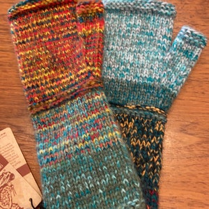Op de afbeelding: Een paar vingerloze handschoenen in een veelkleurig gebreid patroon. E&eacute;n handschoen is een mix van rood, oranje, geel en blauw, terwijl de andere een mix is van blauw, groen en wit.