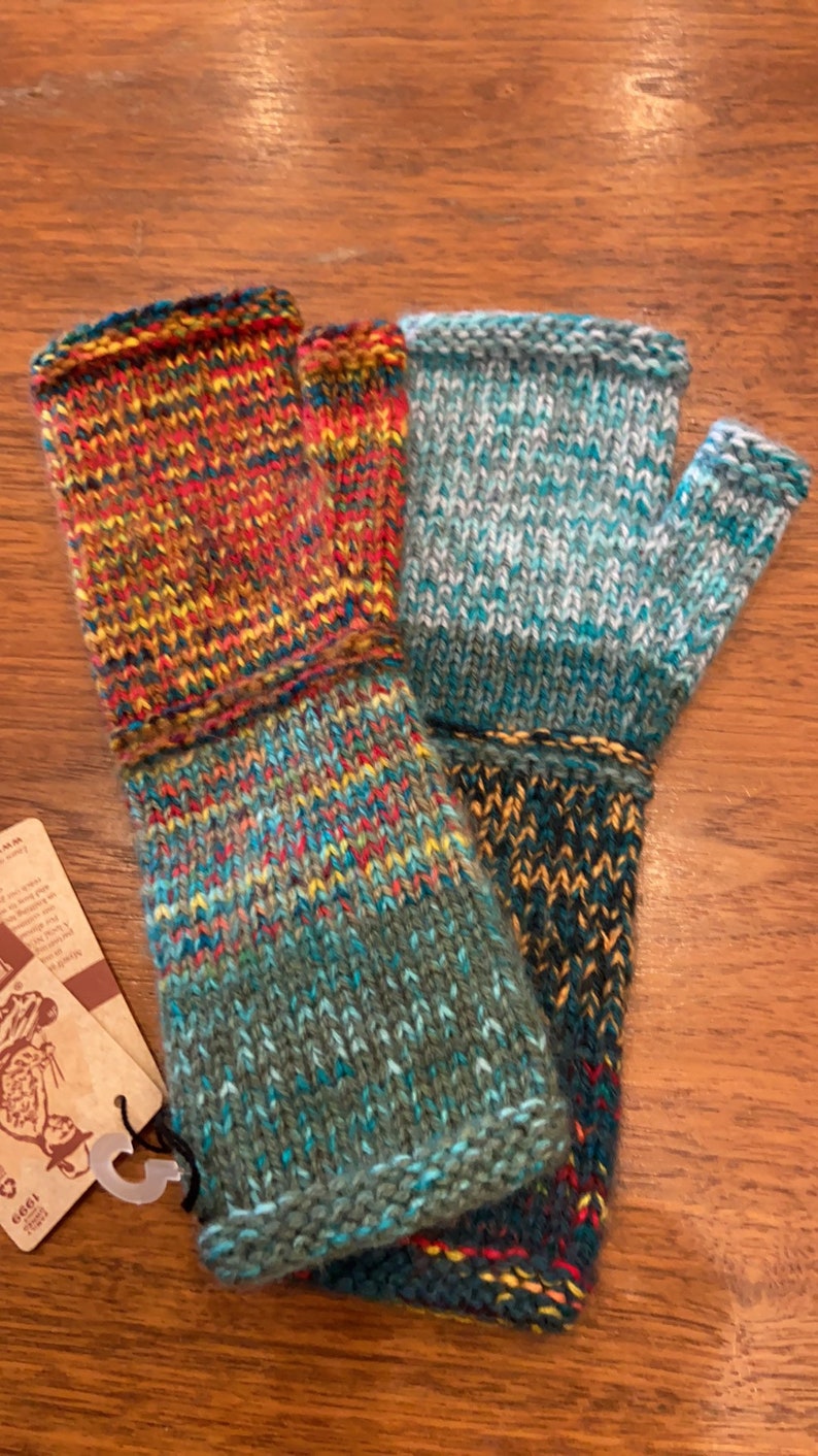Op de afbeelding: Een paar vingerloze handschoenen in een veelkleurig gebreid patroon. E&eacute;n handschoen is een mix van rood, oranje, geel en blauw, terwijl de andere een mix is van blauw, groen en wit.