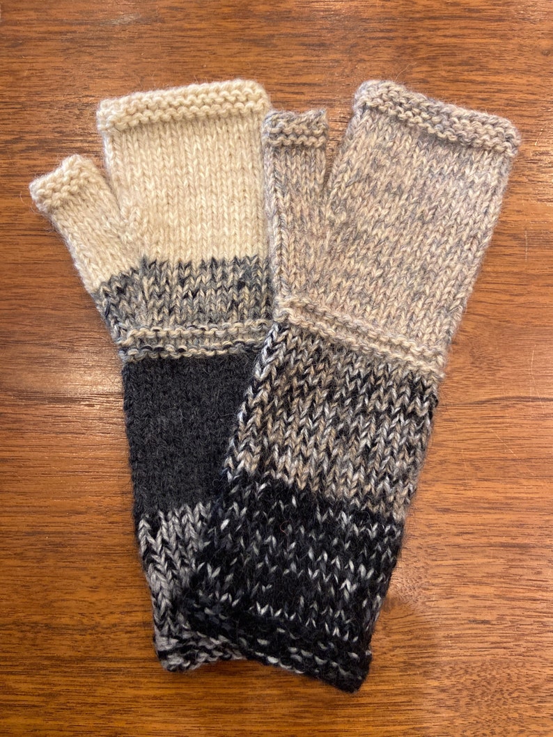 Op de afbeelding: Een paar vingerloze handschoenen met een zwart, grijs en beige breipatroon. De handschoenen zijn gemaakt van een zacht, warm materiaal en perfect om je handen in de winter warm te houden.