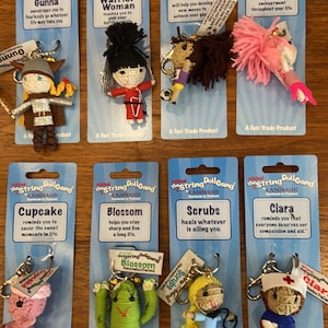 May include: Ten colorful string dolls with keychains, each with a different name and description. The dolls are made of yarn and have different features, such as hair, clothing, and accessories. The dolls are packaged on individual cards with a blue background and white text.