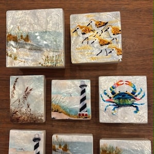 May include: A collection of eight square and rectangular decorative tiles featuring coastal scenes. The designs include a blue crab, lighthouses with black and white stripes, sandy beaches with dunes, wooden fencing, and small shorebirds in shades of tan.