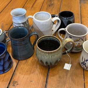 May include: A collection of nine ceramic mugs with various designs and colors. The mugs are handmade and feature unique patterns and glazes.