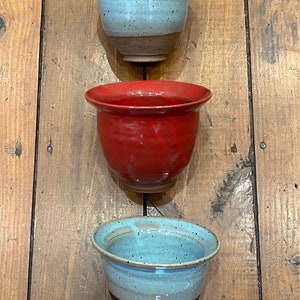 May include: Three ceramic planters in different colors. The top planter is light blue with brown speckles, the middle planter is red, and the bottom planter is light blue with brown speckles and a black and yellow design.