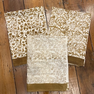 May include: Three folded fabric napkins with a cream background and brown floral patterns. Each napkin has a brown border. The patterns include leaves, flowers, and small sprigs. The napkins are arranged on a wooden surface.