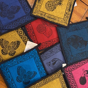 May include: Set of 8 colorful fabric pot holders with various designs, including butterflies, pineapples, fish, lemons, and a fox. The pot holders are square and have black borders.