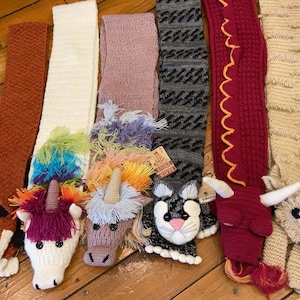 May include: A collection of six long, crocheted animal-themed scarves. The scarves feature designs including a fox, a white unicorn with a rainbow mane, a lavender unicorn, a gray cat, a red dragon with yellow trim, and a beige highland cow.