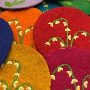 May include: A collection of colorful felt coasters. Each coaster is a different color, including blue, purple, orange, red, yellow, green, and magenta. They are decorated with embroidered white lily of the valley flowers with green stems and leaves.