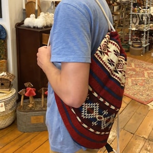 May include: A person wearing a light blue t-shirt and khaki pants is carrying a red, blue, and white drawstring bag with a geometric pattern. The bag has a white drawstring and a black strap.