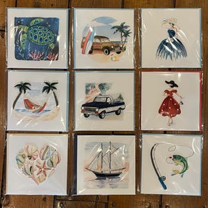 May include: Nine different greeting cards with various summer themes. The cards feature illustrations of a turtle, a car on the beach, a woman in a blue dress, a hammock, a truck, a woman in a red dress, seashells, a sailboat, and a fishing rod with a fish.
