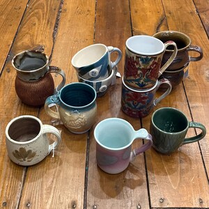 May include: A collection of handmade ceramic mugs in various colors and designs. The mugs feature unique shapes, textures, and patterns, including floral and abstract motifs. Some mugs have a glossy finish, while others have a matte appearance. The mugs are displayed on a wooden surface.