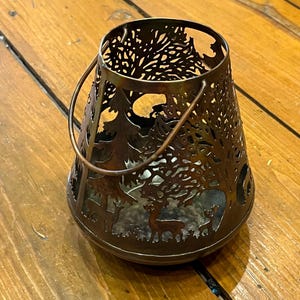 以下が含まれることがあります： 木と鹿がくり抜かれたデザインの茶色の金属製のキャンドルホルダー。ホルダーにはハンドルが付いており、どんな家のインテリアにも素朴な雰囲気を加えるのに最適です。