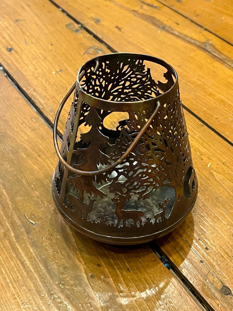以下が含まれることがあります： 木と鹿がくり抜かれたデザインの茶色の金属製のキャンドルホルダー。ホルダーにはハンドルが付いており、どんな家のインテリアにも素朴な雰囲気を加えるのに最適です。